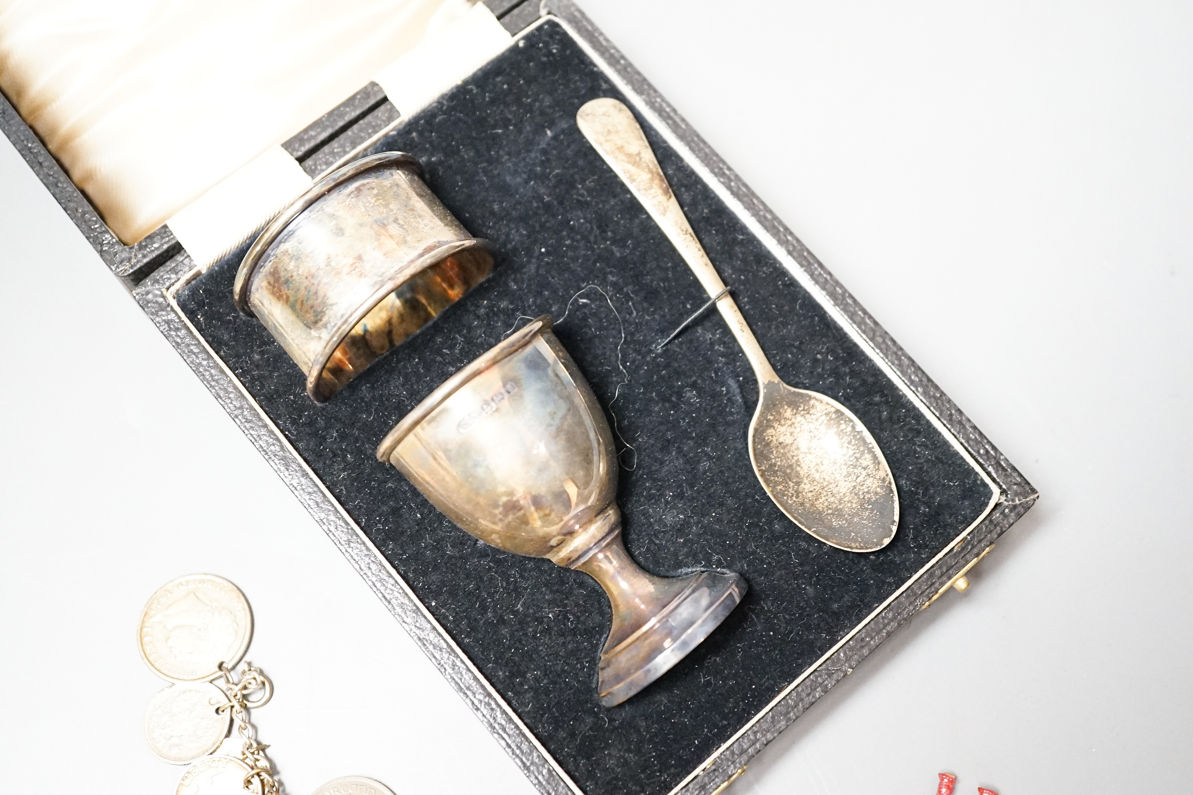 A 1930's cased silver christening set, comprising an egg cup, spoon and napkin ring, together with a white metal charm bracelet hung with assorted charms and coins including silver.